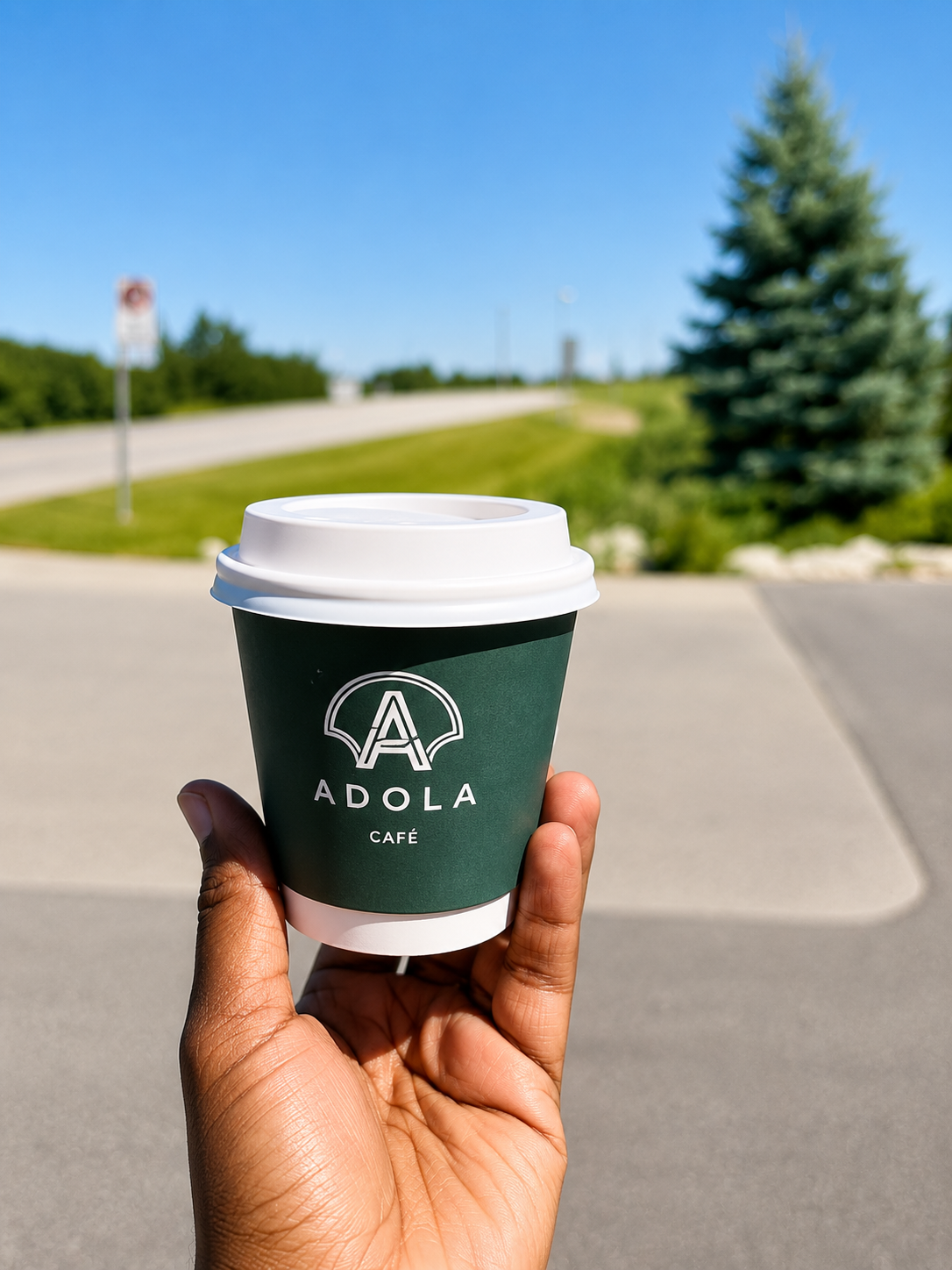 A hand holding an ADOLA Café cup on the open road toward the mountains